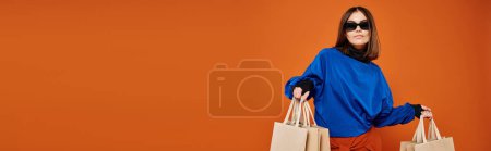 beautiful woman in stylish sunglasses holding shopping bags on orange backdrop, black friday banner