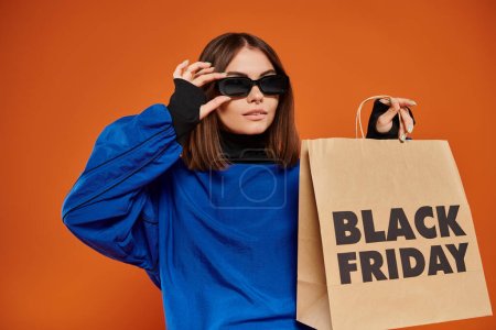 young woman in stylish sunglasses holding shopping bag with black friday letters on orange backdrop