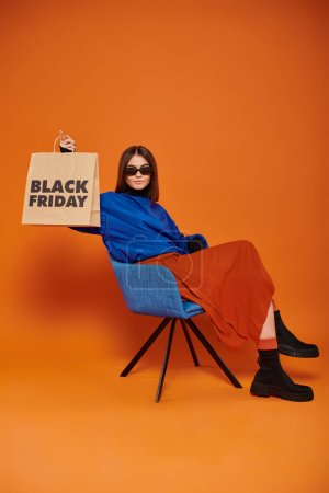Frau in Sonnenbrille und herbstlicher Kleidung mit schwarzer Freitagseinkaufstasche auf Sessel sitzend