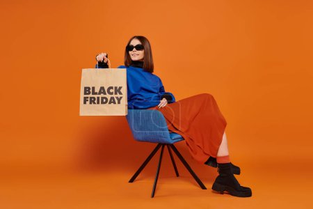 Téléchargez les photos : Femme heureuse dans des lunettes de soleil élégantes tenant sac à provisions noir vendredi et assis sur un fauteuil - en image libre de droit