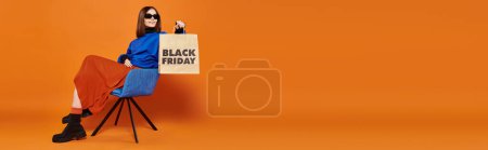 Téléchargez les photos : Femme heureuse dans des lunettes de soleil élégantes tenant sac à provisions noir vendredi et assis sur un fauteuil, bannière - en image libre de droit