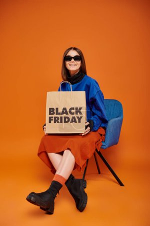 Photo for Cheerful woman in stylish sunglasses holding black friday shopping bag and sitting on armchair - Royalty Free Image