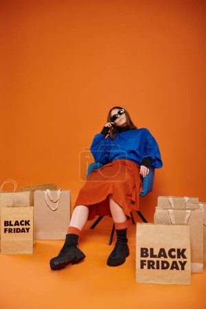 Müde Frau mit stylischer Sonnenbrille sitzt auf einem Sessel neben schwarzen Einkaufstaschen in Orange