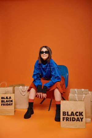 Positive Frau mit stylischer Sonnenbrille sitzt auf Sessel neben schwarzen Freitagseinkaufstüten in Orange