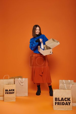 Foto de Mujer feliz sosteniendo caja de cartón con pisos de ballet y de pie cerca de bolsas de compras, viernes negro - Imagen libre de derechos