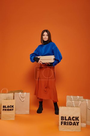 glückliche Frau hält Karton in der Hand und steht neben Einkaufstüten mit schwarzen Freitagsbriefen, orange