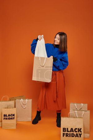 glückliche Frau mit kurzen Haaren hält Einkaufstasche mit weißem Hemd auf orangefarbenem Hintergrund, schwarzer Freitag