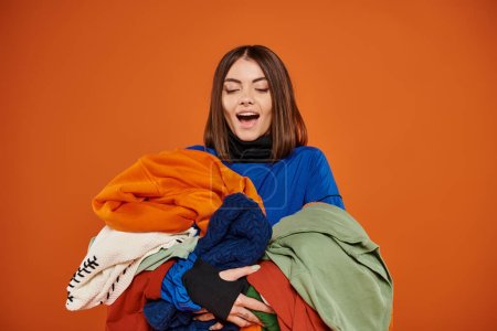 amazed young woman with brunette hair holding pile of colorful autumnal clothes, black friday