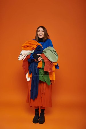 joyous young woman with brunette hair holding pile of colorful autumnal clothes, black friday