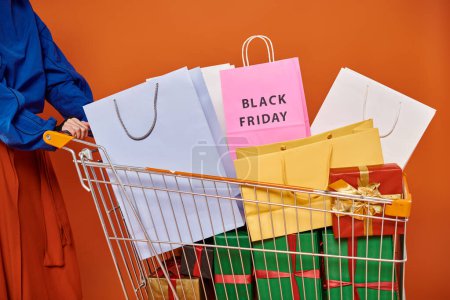 cropped woman standing with cart full of shopping bags, black friday letters on orange backdrop