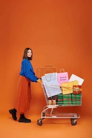 glückliche Frau mit Einkaufswagen voller Einkaufstüten mit schwarzen Freitagsbuchstaben auf orangefarbenem Hintergrund