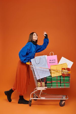 glückliche Frau mit Einkaufswagen voller Einkaufstüten mit schwarzen Freitagsbuchstaben auf orangefarbener, winkender Hand
