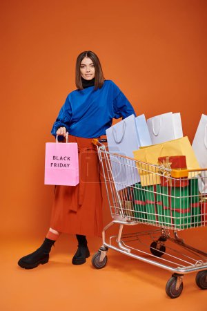 hübsche Frau mit Einkaufswagen voller Einkaufstüten mit schwarzen Freitagsbuchstaben auf orangefarbener, winkender Hand