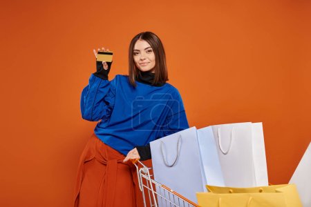 smiling woman holding credit card near cart full of shopping bags on orange, black friday concept