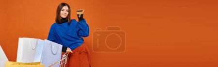 Téléchargez les photos : Femme souriante tenant la carte de crédit près du panier plein de sacs à provisions sur orange, bannière du vendredi noir - en image libre de droit