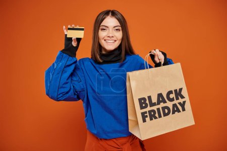 Lächelnde Frau mit Kreditkarte und Einkaufstasche mit schwarzen Freitagsbuchstaben auf orangefarbenem Hintergrund
