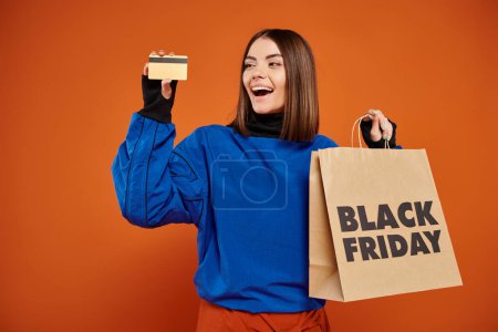 amazed woman holding credit card and shopping bag with black friday letters on orange backdrop