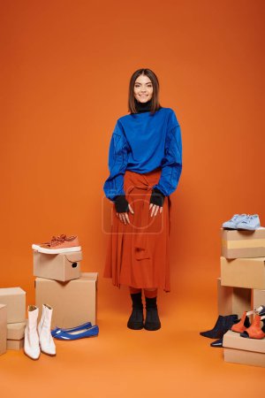 happy woman in autumnal clothes standing near boxes with different shoes on orange, black friday