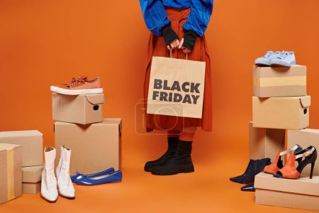 cropped woman in autumnal clothes standing near boxes with different shoes on orange, black friday