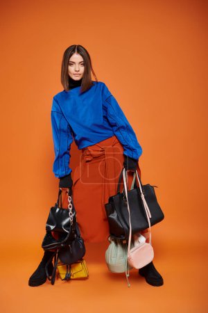 morena mujer en ropa otoñal sosteniendo diferentes bolsas sobre fondo naranja, concepto de viernes negro