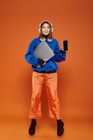 glückliche junge Frau in Kopfhörer und herbstlichem Outfit mit Smartphone und Laptop, Cyber Monday