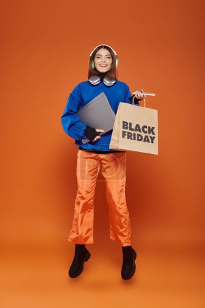 mujer alegre en auriculares y traje otoñal sosteniendo gadgets y bolsa de compras, lunes negro