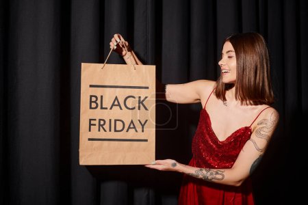 alegre dama sosteniendo bolsa de compras sonriendo felizmente con cortinas negras en el fondo, viernes negro