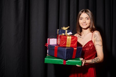 cheerful young woman holding presents and smiling at camera with curtains backdrop, holiday gifts