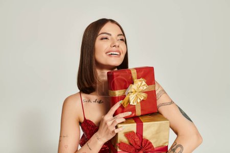 joyous woman with pile of presents in hands looking away on ecru backdrop, holiday gifts concept
