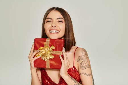fröhliche junge Frau lächelt aufrichtig und hält rotes Geschenk in Händen, Konzept für Weihnachtsgeschenke