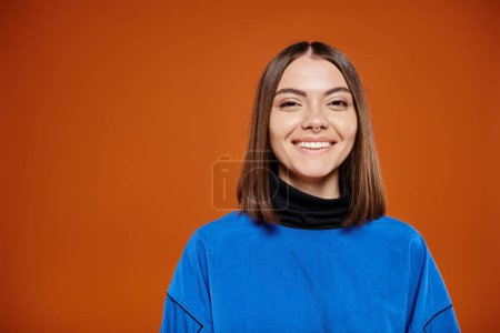 atrakcyjna uśmiechnięta kobieta w luźnej niebieskiej kurtce uśmiechnięta radośnie do kamery na pomarańczowym tle