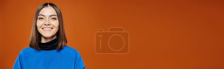 piękna kobieta z przekłutym nosem w luźnej niebieskiej kurtce uśmiechnięta do kamery na pomarańczowym tle, baner