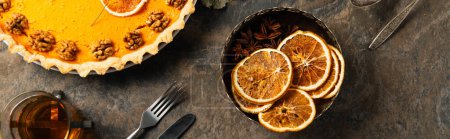 Erntedank-Kürbiskuchen mit Walnüssen neben getrockneten Orangenscheiben und warmem Tee auf Steinoberfläche, Banner