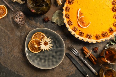 thanksgiving backdrop, decorated pumpkin pie near cutlery, tea and spices with herbs on stone table