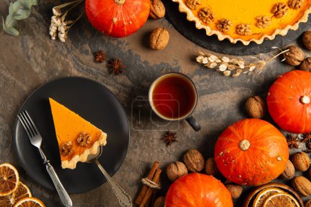 thanksgiving  pumpkin pie on black plate near warm tea and orange pumpkins on decorated stone table