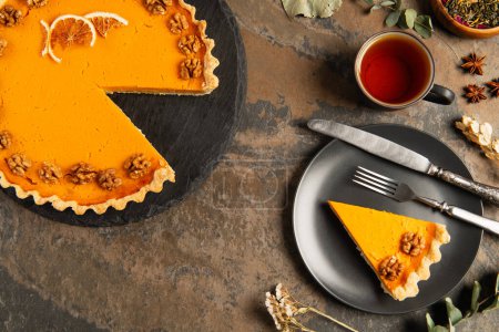 thanksgiving pumpkin pie with walnuts and orange slices near black plate and warm tea on stone table