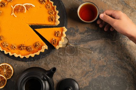 cropped view of man holding cup of warm aromatic tea near thanksgiving pumpkin pie on stone table