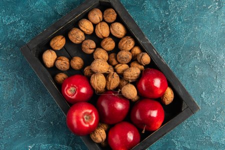 manzanas maduras rojas y nueces enteras en bandeja de madera negra sobre mesa texturizada azul, concepto de acción de gracias
