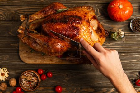 Photo for Cropped view of man cutting thanksgiving turkey near cherry tomatoes and pumpkin on wooden table - Royalty Free Image
