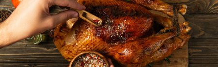 cropped view of man with wooden bowl seasoning grilled turkey for thanksgiving dinner, banner