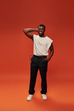joyful african american man in white tank top with hand in pocket of black pants on red, full length