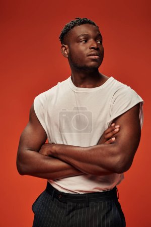 Téléchargez les photos : Jeune homme afro-américain confiant en blanc débardeur regardant loin avec les bras croisés sur rouge - en image libre de droit