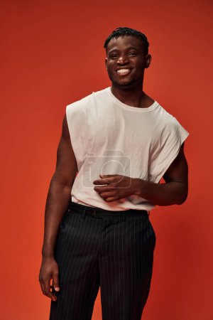 fröhlicher afrikanisch-amerikanischer Typ in weißem Tank-Top und schwarzer Hose, der auf rotem Hintergrund in die Kamera blickt