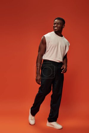 hombre americano africano alegre y caliente en camiseta blanca y pantalones negros mirando hacia otro lado en el fondo rojo