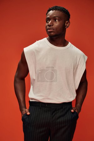 Foto de Modelo masculino afroamericano confiado y elegante cogido de la mano en bolsillos y mirando hacia otro lado en rojo - Imagen libre de derechos
