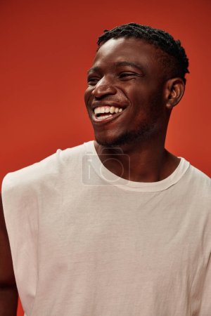 aufgeregter afrikanisch-amerikanischer Mann in weißem Tank-Top, lachend und wegschauend auf rotem Hintergrund, Glück