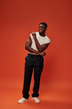 longitud completa de alegre hombre afroamericano en pantalones negros de pie sobre fondo rojo y naranja