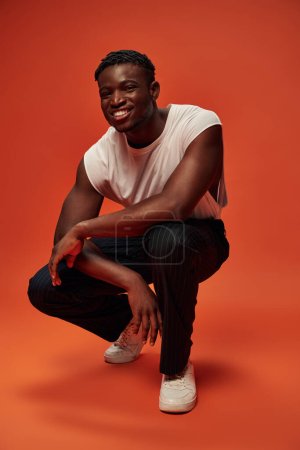 carefree african american man in trendy street wear sitting on haunches on red and orange backdrop
