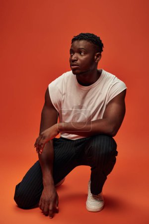serious african american man in white tank top sitting on haunches and looking away on red backdrop