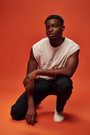 Photo for Self-confident african american man in trendy casual clothes sitting on haunches on red, full length - Royalty Free Image